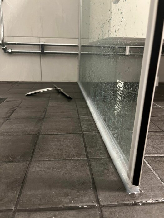 Ett duschhörn med glasvägg, grå klinkergolv, vattendroppar på glaset, en del av en duschskrapa synlig.