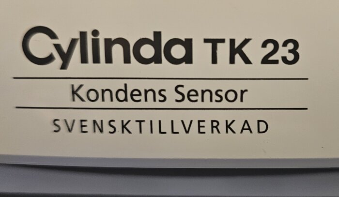 Etikett med text "Cylinda TK 23 Kondens Sensor SVENSKTILLVERKAD", svart på vit bakgrund.