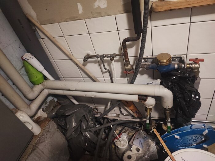 Rörledningar och vattenmätare med diverse föremål i oordnad miljö, eventuellt under renovering eller reparation.