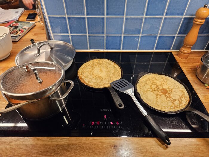 Två pannkakor på stekjärn, kokkärl på spishäll, blå kakelvägg i bakgrunden, träpepparkvarn, matlagning i hemmiljö.