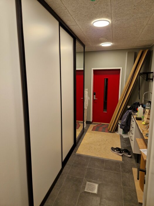 Inomhusmiljö med röda dörrar, byggmaterial, mattor, skor och en glasskjutdörr. Modern belysning och skrivbordsmaterial synligt.