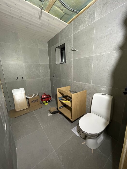 Under renovering, badrum med toalett och osammanbyggd inredning, grå kakel, verktyg och byggmaterial på golvet.