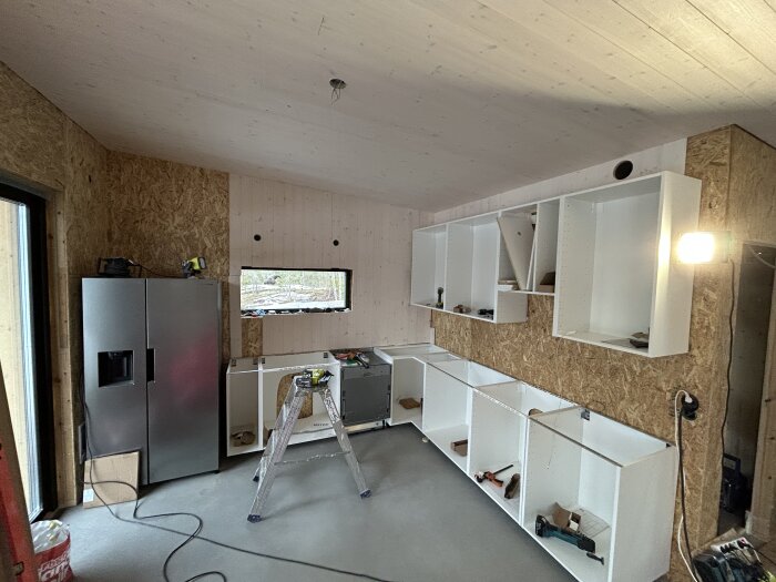 Interiör av ett rum under konstruktion med oskyddade väggar, installation av skåp och kylskåp, stege och verktyg synliga.