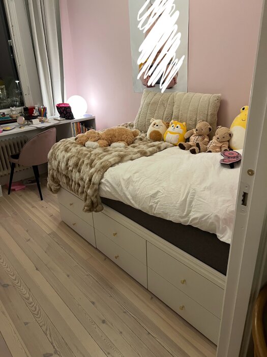 Mysigt sovrum med säng, mjukisdjur och skrivbord, rosa väggar och trämönstrat golv.