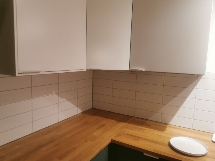 Hörn av kök med vita skåp, kakelvägg, träbänkskiva och en tom tallrik.
