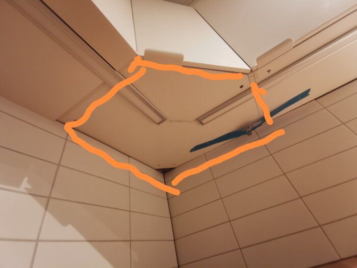 Taket i ett rum med vita plattor, ventilationsgaller, en blå detalj och orangea markeringar.