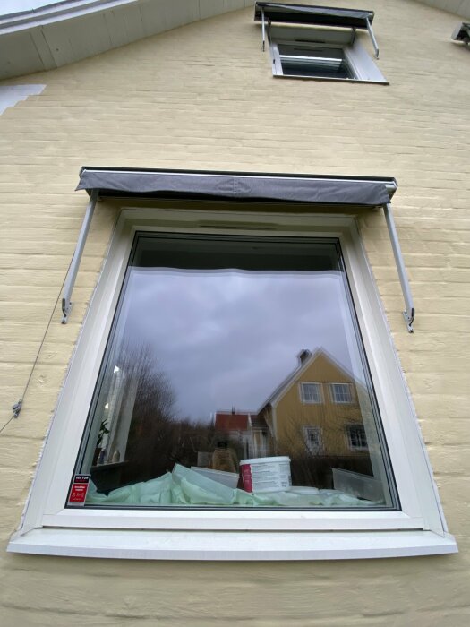 Ett öppet fönster i ett gult hus, reflektion av en molnig himmel, fönstermarkis ovanför.