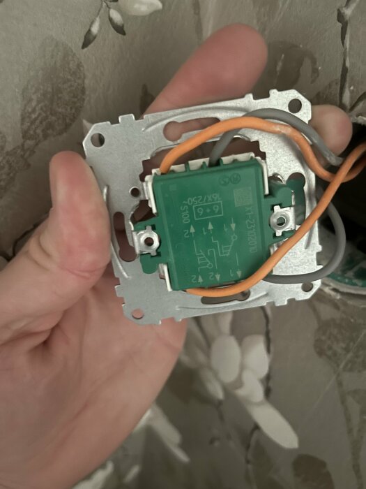 Hand håller en öppen elektrisk väggkontakt med synliga ledningar och elektroniska komponenter under installation eller reparation.