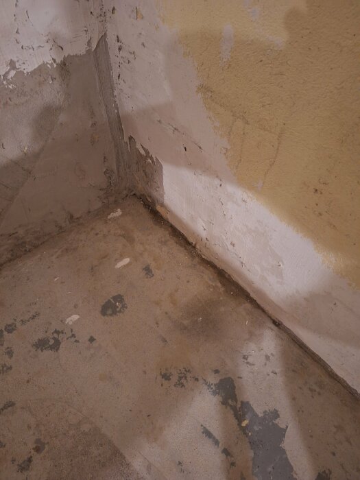 Ett odetaljerat hörn med fläckigt golv och avflagande väggfärg. Indikerar renoveringsbehov eller vattenskada.