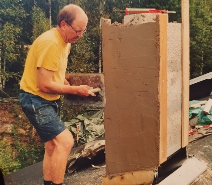 Man applicerar cement på en vertikal yta utomhus med grönska i bakgrunden. Arbetskläder och verktyg syns.