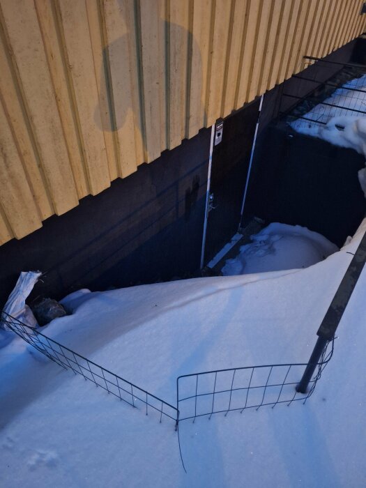 Nedgång täckt av snö vid kväll, vid en gul byggnad, med staket och varningsskylt.