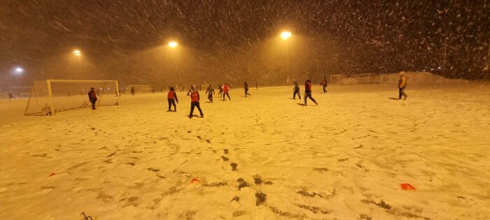 Personer spelar fotboll på snötäckt plan i kraftigt snöfall under belysning på kvällen.