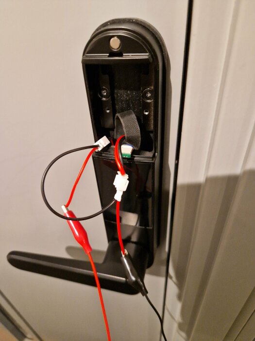 Ett batterigreb med kablar anslutna, möjlig provisorisk reparation eller testning, mot vit dörr.