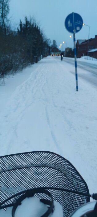 Snötäckt gång- och cykelväg, skymning, trafikljus på avstånd, del av cykelkorg i förgrunden.