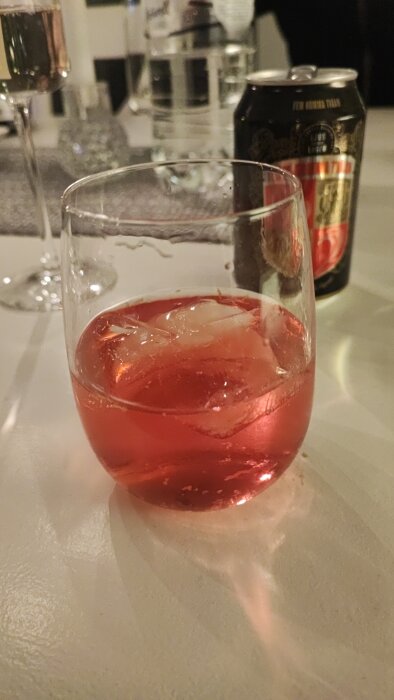 Ett glas med röd dryck och is, suddig bakgrund, dryckesburk, vattenglas, festlig stämning.