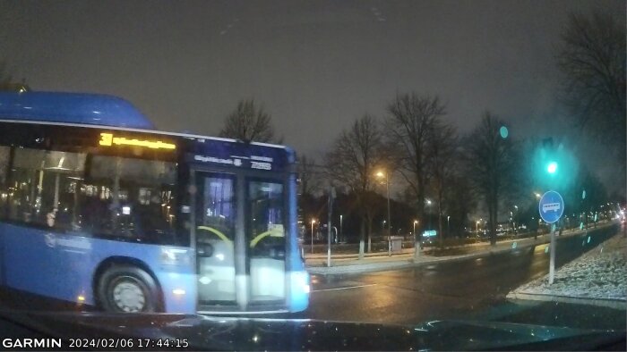 En blå buss körs i kvällstrafik vid ett trafikljus som lyser grönt. Längs en våt väg med träd.