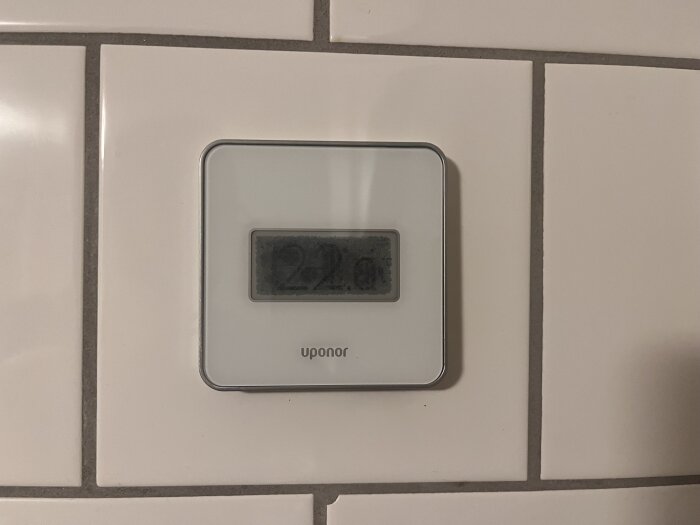 Vit termostat på kakelvägg visar 29 grader, märkt med "Uponor".