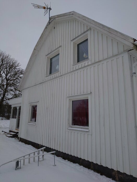 Ett vitt hus i snöigt landskap med tv-antenn och metallhandräcke. Korrugerad sidbeklädnad och fönsterreflektioner.