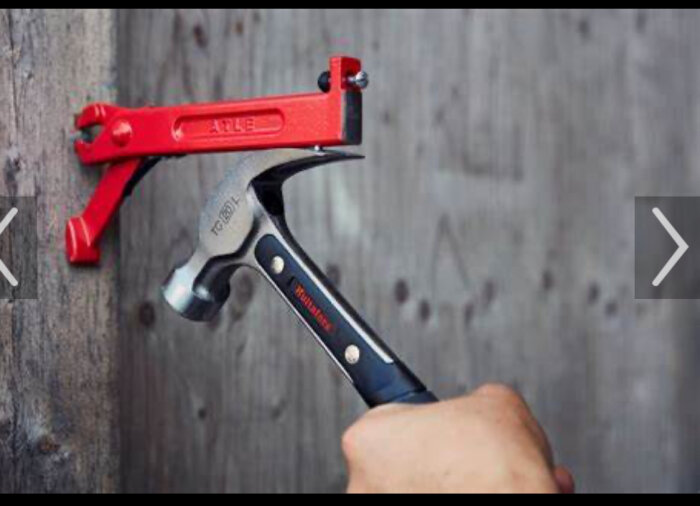 En hand håller en hammare nära en spik på grått trä. Röd slagklots hänger bredvid.