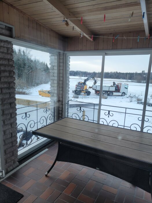 Utsikt från inglasad terrass med bord över vinterlandskap, anläggningsfordon och snöklädda träd i bakgrunden.