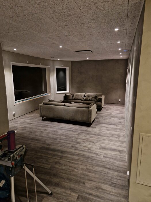 Modernt vardagsrum under renovering, soffa framför inbyggd TV, trägolv, infällda spotlights, arbetsmaterial i förgrund.