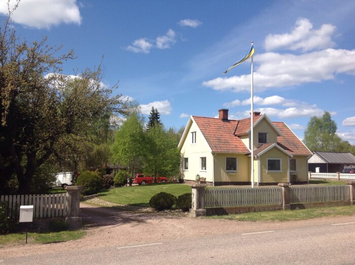 Ett gult hus med rött tak, svensk flagga, grönområde, staket, klar himmel och en bil.