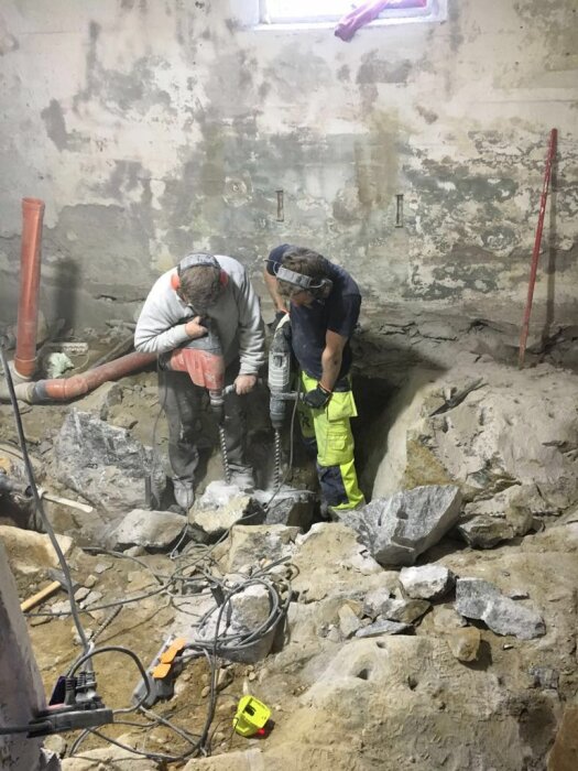 Två arbetare i säkerhetskläder använder verktyg för att bryta upp betong under en renovering inomhus.