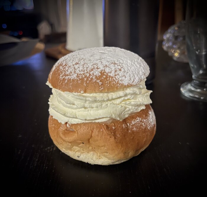 Semla med fluffig grädde, pudrad med florsocker, på mörk yta.