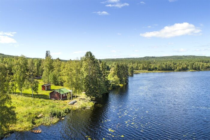 Sjöstrand med röda stugor, gröna träd, klarblå himmel och lugnt vatten. Idyllisk natur, troligen Skandinavien.