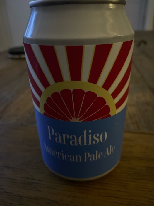 Aluminiumburk med grafisk design, röda strålar, "Paradiso American Pale Ale", dryck, träyta, inomhusljus.