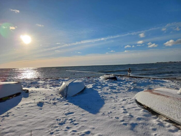 Vintersol över snötäckt strand med båtar och spegelblankt hav.