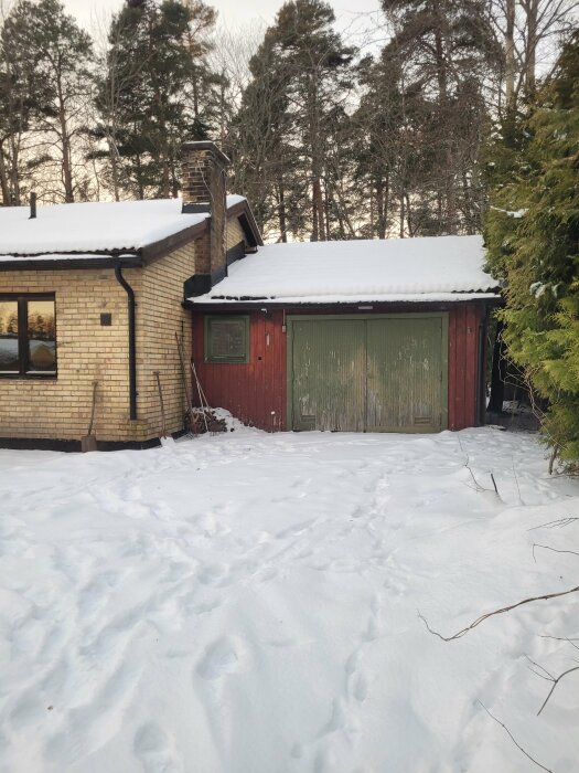 Hus med snö på taket, grön garageport, spår i snön, träd i bakgrunden, vinterdag.