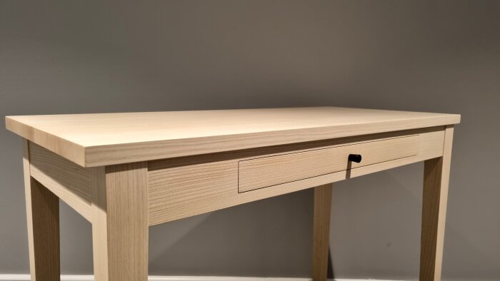 Ljust träskrivbord med en låda, minimalistisk design, mot grå vägg, modern inredning.