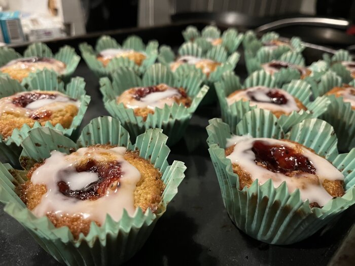 Nybakade muffins med glasyr och sylt i gröna formar på en mörk yta.