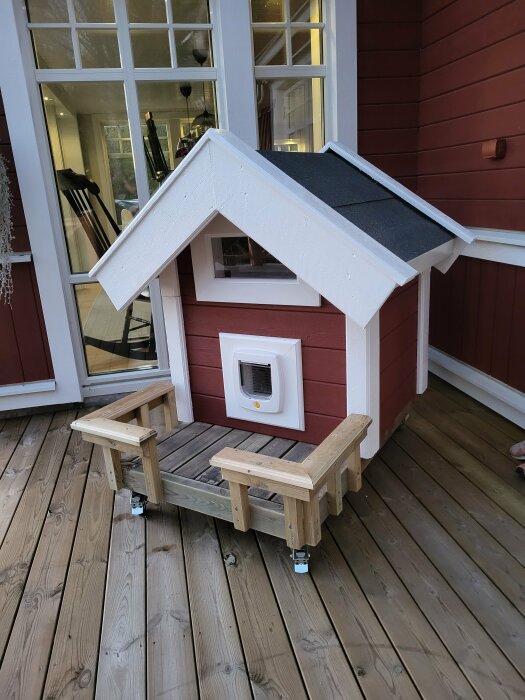 Litet, rött och vitt hundhus med veranda på trädäck utanför ett stort hus.