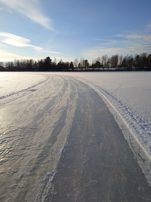 Isbelagd sjö, vintersol, spår i snö, träd i bakgrunden, klar himmel.