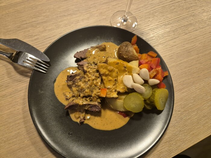 Middagstallrik med stekt kött, sås, potatis, röd paprika, pickles, vitlök på träbord.