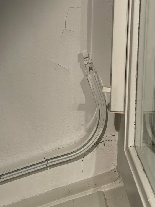 Vit vägg med kablar i kabelkanal nära ett fönster och sprickor i väggens målning.