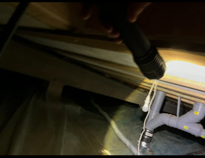 En handlampe lyser under en säng eller möbel, avslöjar rörledningar och konstruktionen nedan.