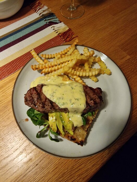 En måltid med stekt kött, sås, avokado, potatisgaller (crinkle cut fries) på en tallrik, träbord i bakgrunden.