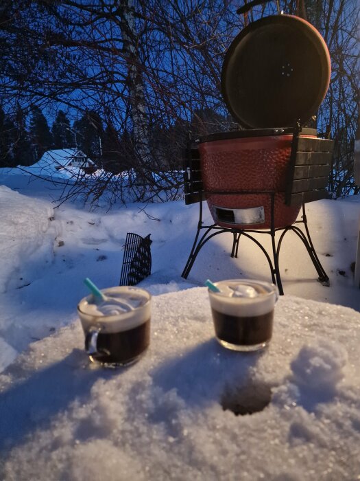 Två kaffedrinkar på ett snöigt bord, grill i bakgrunden, skymning, snöig vintermiljö, utomhus, avkopplande stämning.