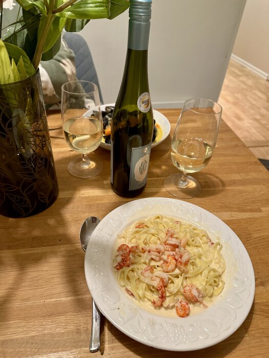 Middagsdukning med pasta, räkor, vitvin, blommor och en delvis synlig person i bakgrunden.