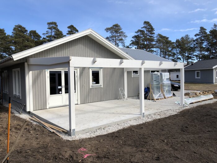 Ett modernt, nybyggt enplanshus med terrass, fönster och byggmaterial runtomkring under en klar himmel.