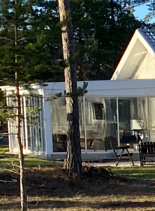 Träd framför hus med vita väggar, stora fönster och ett rött tak, omgivet av natur.