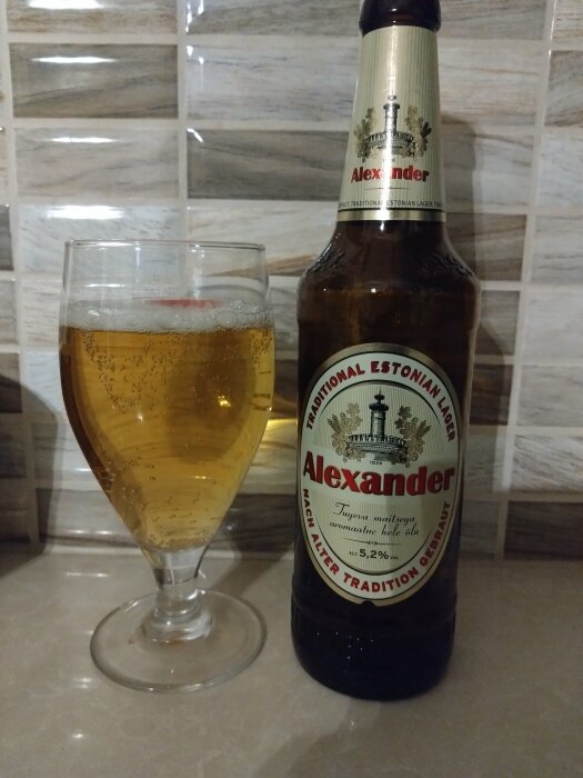 En ölflaska av märket Alexander och ett glas med öl framför kakelvägg.