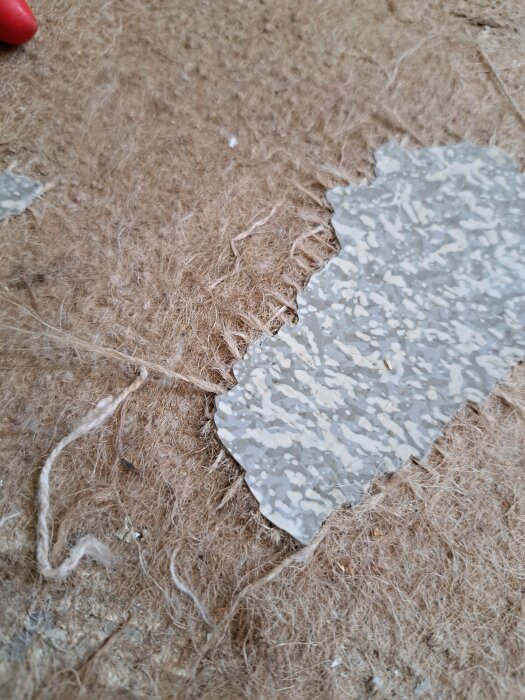 Närbild av grov textil med en bit flagnad, mönstrad färg eller material. Slitna och fransiga fibrer syns.