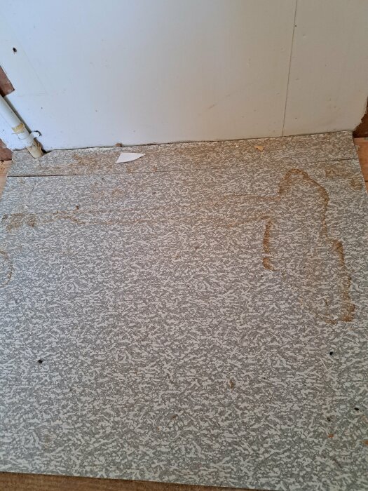 Gammalt grått linoleumgolv med vattenskada och spår av slitage nära vit vägg.