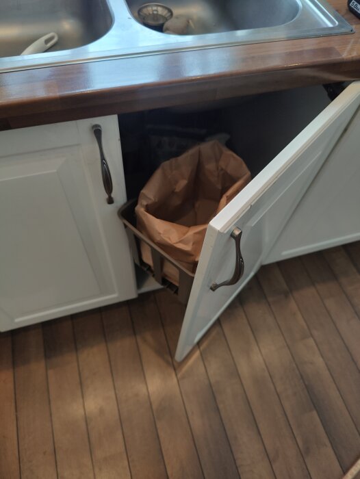 Köksunderskåp öppet med en brun papperspåse, diskbänk ovanför, träarbetsytor, och handtag i metall.