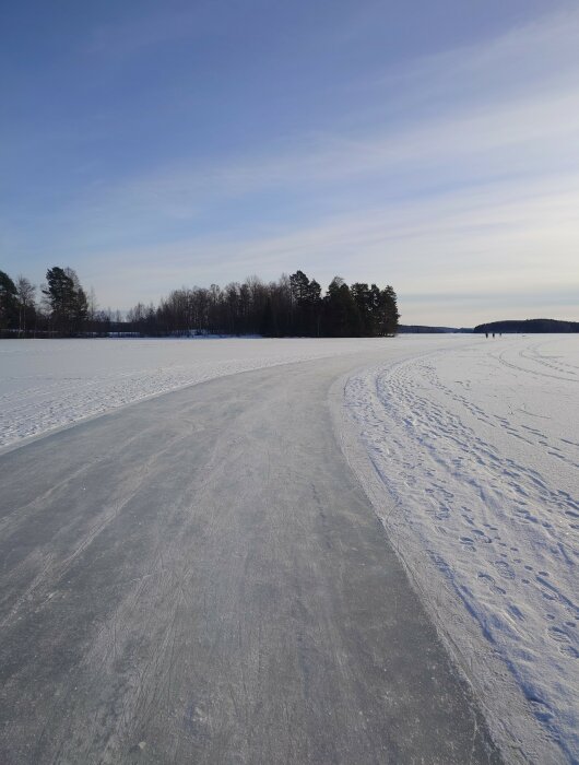 Istäckt sjö med snö, blå himmel, skog i horisonten, plogad skridskobana.