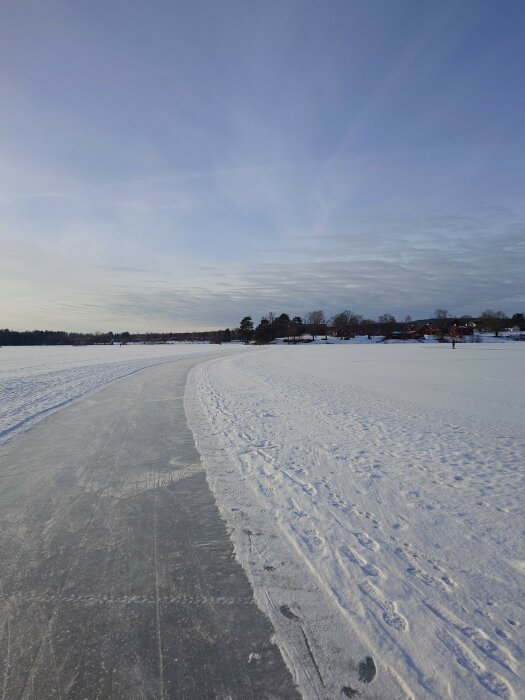 Istäckt sjö, upplogad skridskobana, vintersol, skogsbryn, blå himmel, snödrivor, fotspår.